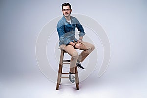 Full length portrait of an attractive young man in jeans shirt sitting on the chair over grey background.