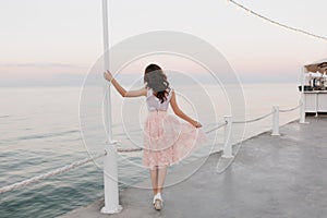 Full-length portrait from back of elegant brunette girl walking along the ocean wharf and enjoying beautiful morning