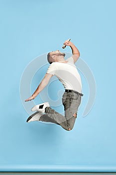 Full-length portrait of bearded energetic man in casual white t-shirt singing in microphone, jumping against blue studio