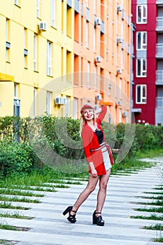 Full length portrait of beautiful blonde young hipster woman in red blouse and cap posing near bright buildings