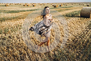 Full length portrait of a beautiful brunette in a dress and with a warm plaid. Woman enjoying a walk in a wheat field with hay