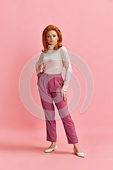 Full-length portrait of beautiful redhead woman in stylish clothes, hairdo and makeup posing against pink studio