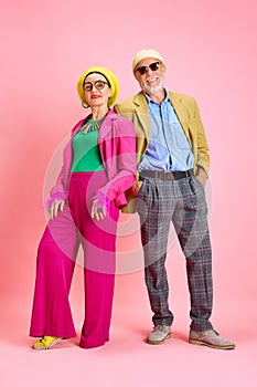 Full-length portrait of beautiful senior couple, elegant man and woman in stylish clothes standing against pink