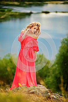 Full length portrait of beautiful sensual young blond woman in long pink dress and floral wreath near the lake outdoors