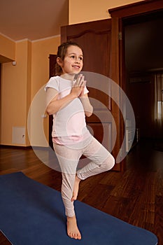 Full length portrait of beautiful smiling girl child practicing yoga indoors, standing in tree pose on blue fitness mat