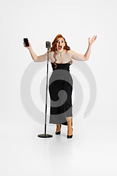 Full-length portrait of beautiful woman in stylish black dress holding phone isolated over white background. Excited