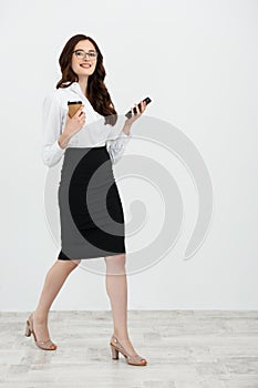 Full length Portrait of beautiful young business woman in formal wear walking and texting on cell phone with takeaway