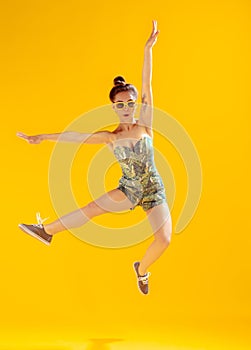 Full-length portrait of beautiful young girl posing, cheerfully jumping isolated over yellow studio background. Having