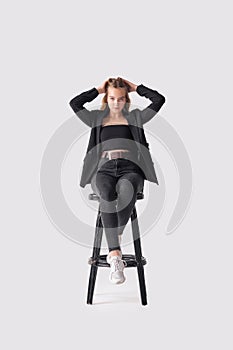 Full length portrait of beautiful young girl sitting on tall chair in black jeans and jacket over gray background