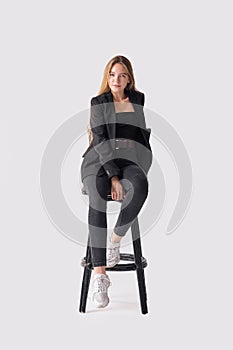 Full length portrait of beautiful young girl sitting on tall chair in black jeans and jacket over gray background