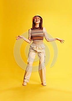 Full-length portrait of beautiful young girl in stylish outfit with panama and glasses, posing, dancing against yellow