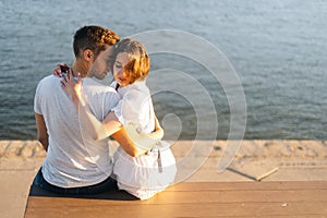 Full length portrait of beautiful young happy couple in love hugging, sitting on bench on city waterfront near river in