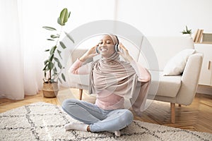 Full length portrait of black Muslim woman in hijab and headphones listening to music on floor in living room