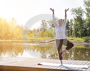 Full-length portrait of a Caucasian elderly woman in yoga vrikshasana poses, tree pose, against the background of a lake