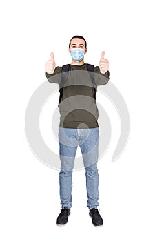 Full length portrait of cautious young man wearing face mask as protection against coronavirus, shows both thumbs up isolated on