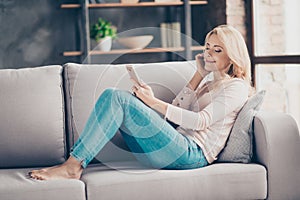 Full length portrait of charming pretty woman in jeans and pullover siting with legs on couch in livingroom, having earphones, us