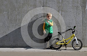 Full length portrait of cheerful little child boy with yellow bicycle standing near concrete wall and pointing by finger on space