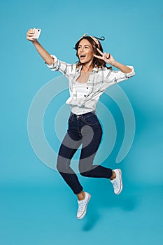 Full length portrait of cheerful woman 20s wearing headband smiling and showing peace sign while taking selfie on mobile phone, i