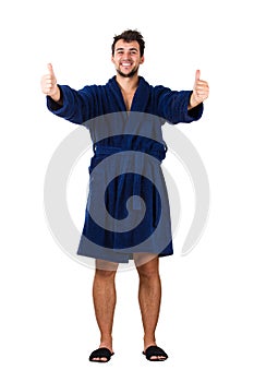 Full length portrait of cheerful young man, positive smiling showing thumbs up gesture, wears blue bathrobe, isolated over white