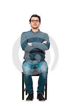 Full length portrait of confident businessman seated on a chair, wearing eyeglasses and keeps arms crossed, isolated on white
