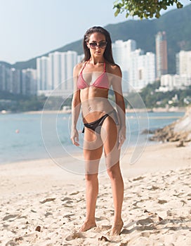 Full-length portrait of confident fitness female model wearing swimsuit standing on sandy beach with high buildings in