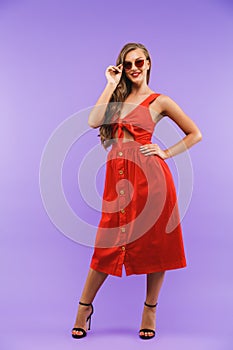 Full length portrait of content pretty woman 20s wearing red dress smiling at camera, standing isolated over violet background