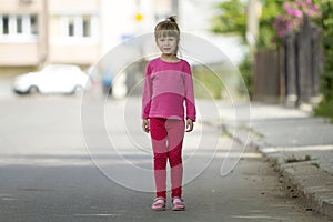 Full length portrait of cute small girl in pink casual clothing and long blond pony tail looking in camera standing on blurred bri