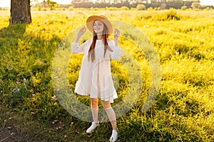 Full length portrait of cute smiling redhead woman in straw hat and white dress standing posing with closed eyes on