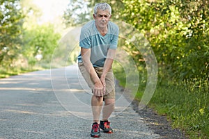 Full length portrait of displeased male pensioner, leans on knees, has pain after jogging, banged his leg, stands on road in rural photo