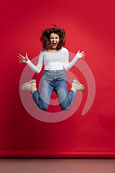 Full-length portrait of excited young beautiful girl in casual clothes jumping isolated on red color background