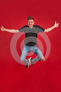 Full length portrait of an excited young man in white t-shirt jumping while celebrating success isolated over red background.