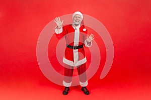 Full length friendly smiling gray bearded man in santa claus costume raising hands greeting and welcoming looking at camera