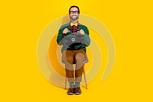 Full length portrait of funky positive man sit chair hands hold wheel isolated on yellow color background