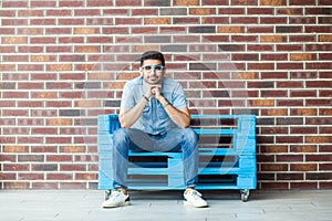 Full length portrait of funny happy handsome young bearded man in blue shirt and eyeglasses sitting on blue wooden pallet and