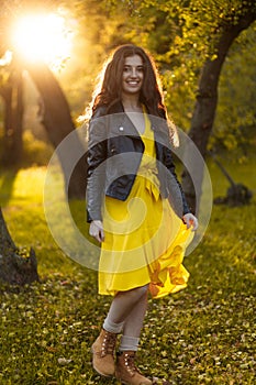 Full length portrait of a girl with long hair, in a jacket and dress in the park,, against the background of the sunset