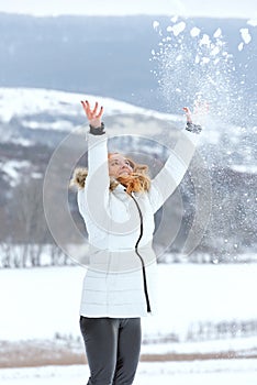 Full-length portrait of girl in white down jacket tossing snow. Winter smiling woman. Vertical frame