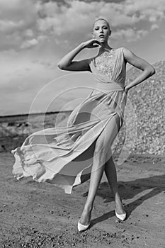 Full length portrait of a gorgeous young woman in flying dress poses in sand quarry on sunny day, clouds background