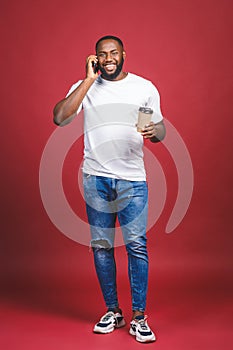 Full length portrait of handsome African American man with mobile phone and take away coffee cup. Isolated over red background