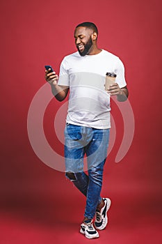 Full length portrait of handsome African American man with mobile phone and take away coffee cup. Isolated over red background