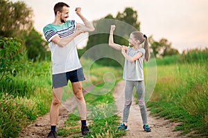 Full length portrait of handsome father and little daughter showing their muscles, looking at camera and smiling outdoor