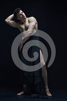 Full-length portrait of handsome man with relief, muscular, shirtless body posing in dark fabric against black studio