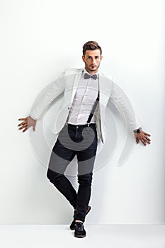 Full length portrait of handsome man in white elegant suit posing and leaning at wall