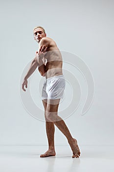 Full-length portrait of handsome young man with blonde hair, muscular body posing in underwear against grey studio