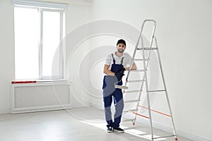 Full length portrait of handyman with roller brush near ladder indoors. Professional construction tools