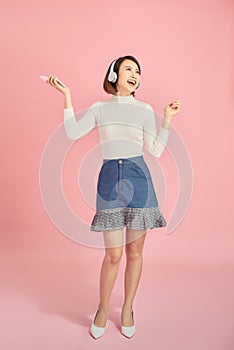 Full length portrait of a happy asian businesswoman listening to music with headphones while dancing isolated over pink background
