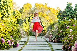 Full length portrait of happy barefoot attractive woman in stylish red white dress holding shoes and walking on tile path in