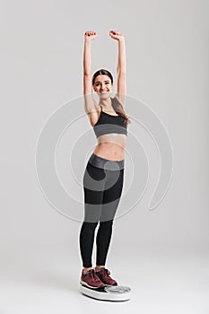 Full-length portrait of happy gymnastic woman measuring her weight after fitness standing on scale, isolated over gray background