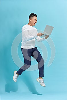 Full length portrait of happy man jumping and holding  laptop isolated over blue background