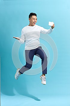 Full length portrait of happy man jumping and screaming while taking selfie on smartphone over blue background