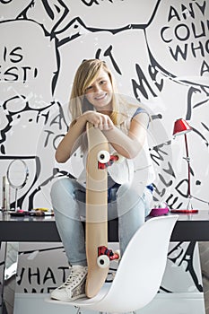 Full-length portrait of happy teenage girl with skateboard sitting on study table at home